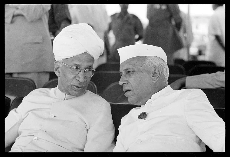 Black-and-white photograph of Jawaharlal Nehru in a white cap leaning toward another seated Indian official wearing a turban