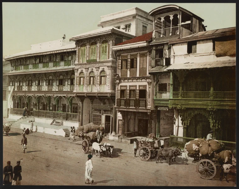 Colour photochrom of a crowded Bombay street around 1890, lined with balconied buildings, shopfronts, awnings, and pedestrians