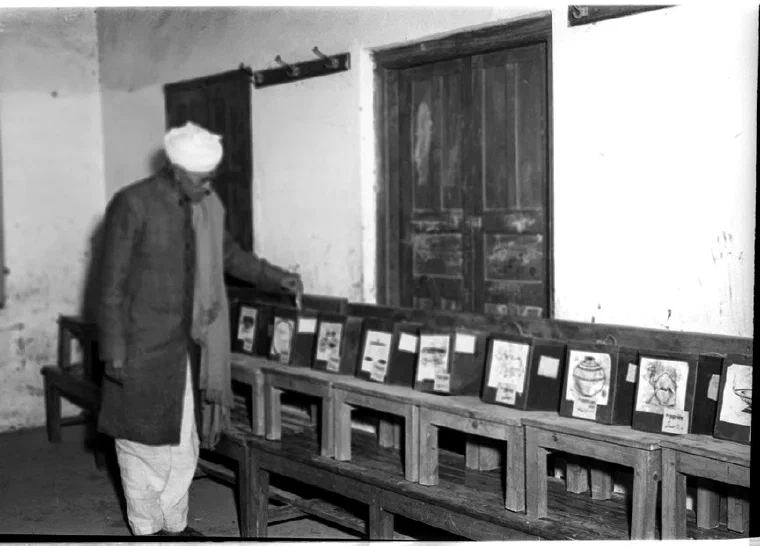 Black-and-white photograph of an Indian villager casting a ballot behind a curtained booth in Delhi in 1952